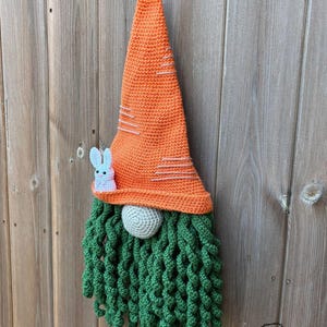 May include: A handmade crochet gnome with an orange carrot-shaped hat and a green beard. A small, light blue bunny is attached to the hat. The gnome's nose is a gray ball. The gnome is a decorative item.