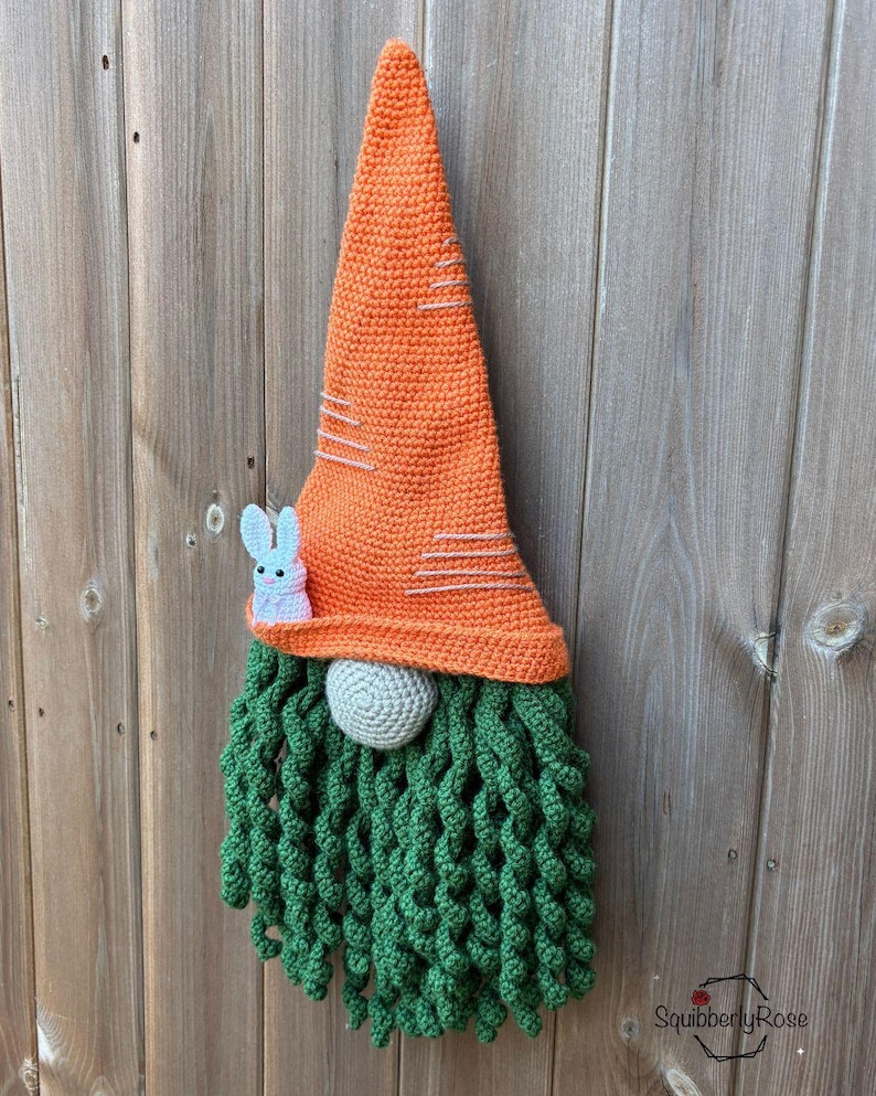 May include: A handmade crochet gnome with an orange carrot-shaped hat and a green beard. A small, light blue bunny is attached to the hat. The gnome's nose is a gray ball. The gnome is a decorative item.