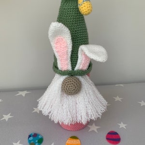 May include: A crocheted Easter gnome with a green hat, pink bunny ears, and a white beard. The gnome is holding a yellow Easter egg and is surrounded by three colorful Easter eggs.