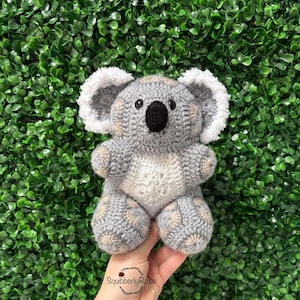 Puede incluir: Un peluche de koala hecho a mano con ganchillo. El koala es principalmente gris con detalles blancos en las orejas y el vientre. El oso tiene una nariz negra y ojos negros pequeños. El peluche se sostiene sobre un fondo verde.