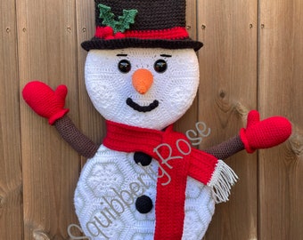 May include: A crocheted snowman with a black top hat, red scarf, and red mittens. The snowman has a white body with black eyes, a carrot nose, and a smiling mouth.