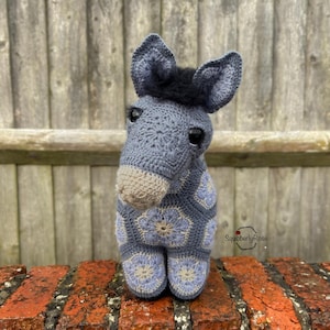 May include: A crocheted gray donkey with a black mane and large brown eyes. The donkey is standing on a brick surface and has a floral pattern on its body. The text "SquirrellyRose" is visible in the lower right corner of the image.