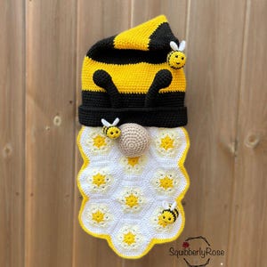 Op de afbeelding: Een handgemaakte gehaakte kabouter met een bijen thema. De kabouter heeft een geel en zwart gestreepte hoed, zwarte antennes en een wit en geel honingraatvormig lichaam. Drie kleine bijen applicaties zijn bevestigd. De tekst "SquibberlyRose" staat rechtsonder.