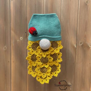 Op de afbeelding: Een handgemaakte gehaakte kabouter met een blauwgroene hoed, een rode lieveheersbeestje en een witte balneus. De baard van de kabouter is gemaakt van gele en bruine gehaakte zonnebloemen. De kabouter wordt getoond tegen een houten achtergrond.