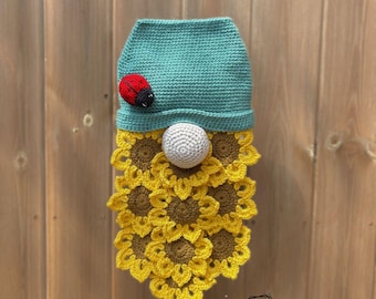 Op de afbeelding: Een handgemaakte gehaakte kabouter met een blauwgroene hoed, een rode lieveheersbeestje en een witte balneus. De baard van de kabouter is gemaakt van gele en bruine gehaakte zonnebloemen. De kabouter wordt getoond tegen een houten achtergrond.
