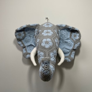 Peut inclure: Une tête d'éléphant en crochet à suspendre au mur, en gris et bleu clair avec des défenses blanches.