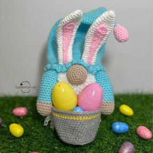 May include: A crocheted gnome wearing a blue hat with white and pink bunny ears, holding a grey basket filled with a yellow and a pink plastic egg. The gnome has a tan nose and white beard. The text SquibberlyRose is visible on the left.