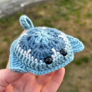 May include: A small, crocheted stingray plush toy crafted from variegated blue and white yarn. The creature features a rounded body, two side fins, a small top loop, and two shiny, round black plastic eyes set into the textured surface.