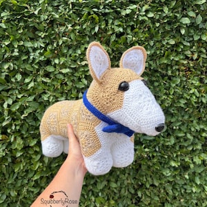 Può includere: Un cane corgi all'uncinetto fatto a mano, con filato beige e bianco. Il cane ha muso, orecchie e zampe bianche, con occhi neri e un collare blu. Il corgi è tenuto davanti a uno sfondo di foglie verdi.