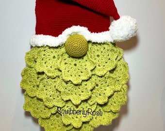 May include: A green crocheted gnome with a red crocheted Santa hat and a white pom-pom. The gnome's beard is made of crocheted green circles. The gnome's nose is a small crocheted brown ball. The gnome's eyes are two small black buttons. The gnome's hat has a white crocheted trim. The gnome is hanging on a white wall. The text "SquiberblyRose" is visible on the gnome's beard.