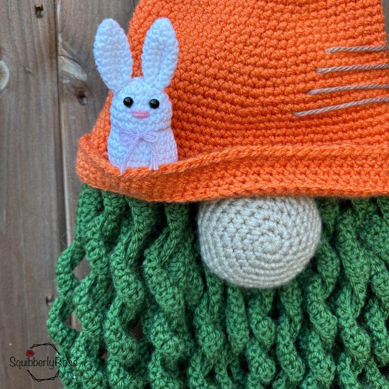 May include: A handmade crochet gnome with an orange hat, green beard, and a white bunny accent. The gnome's hat has a stitched design. The bunny has a pink bow. The gnome's nose is a light beige color.