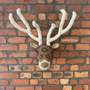 May include: A crocheted deer head with brown and white antlers and a brown and white face with floral details. The deer head is mounted on a brick wall.