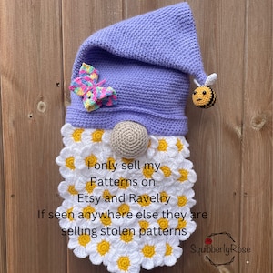 May include: A crocheted gnome with a purple hat, a white daisy beard, and a bee charm. The gnome is holding a colorful butterfly charm. The text "I only sell my Patterns on Etsy and Ravelry If seen anywhere else they are selling stolen patterns Squibberly Rose" is written below the gnome.