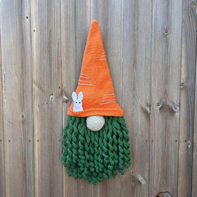 May include: A handmade Easter gnome decoration with a carrot-shaped orange hat and a green beard. The hat has white stripes and a small white bunny applique. The gnome has a round, cream-colored nose.