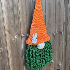 May include: A handmade crochet gnome with an orange carrot-shaped hat and a green beard. The hat features white stripes and a small white bunny applique. The gnome's nose is a light gray sphere, perfect for Easter decor.