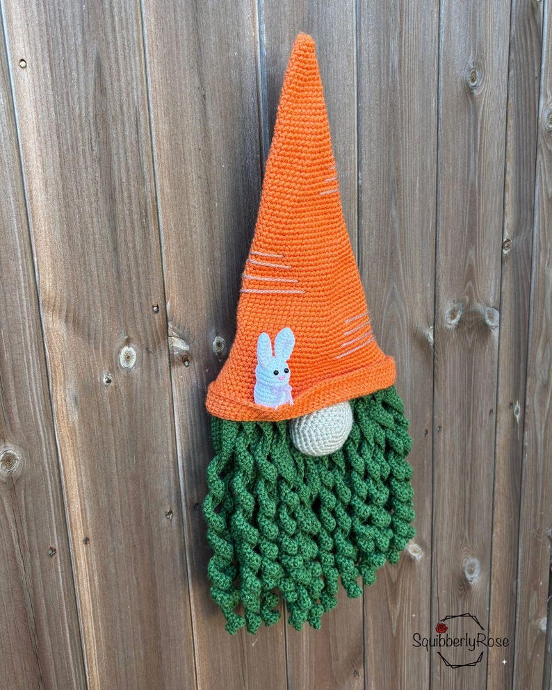 May include: A handmade crochet gnome with an orange carrot-shaped hat and a green beard. The hat features white stripes and a small white bunny applique. The gnome's nose is a light gray sphere, perfect for Easter decor.
