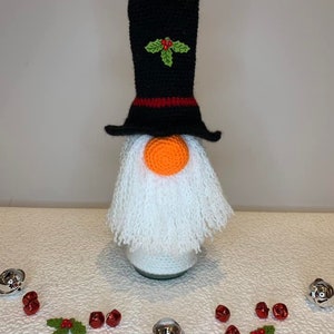 Puede incluir: Un tapón de botella de gnomo de ganchillo con un sombrero de copa negro, barba blanca y nariz naranja. El sombrero tiene una banda roja y una rama de acebo verde. El gnomo está rodeado de cascabeles plateados y rojos.