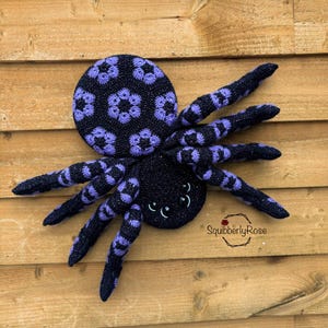 Puede incluir: Una araña de crochet negra y morada con un patrón de flores en la espalda y grandes ojos negros. La araña está sentada sobre una superficie de madera.