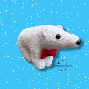 May include: A handmade white crochet polar bear with black eyes and a red bow tie. The bear is set against a light blue background with white snowflake accents. The bear is a decorative item.