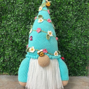 May include: A handmade crochet gnome with a turquoise hat decorated with white and pink flowers, green leaves, and a bee. The gnome has a white beard, a tan nose, and a turquoise body. This decorative item is handmade.