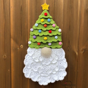 May include: A handmade crochet Christmas gnome decoration. The gnome features a green tree-shaped hat adorned with colorful pom-poms and a yellow star. The beard is white, and the nose is a beige sphere. The gnome is displayed against a wooden background.