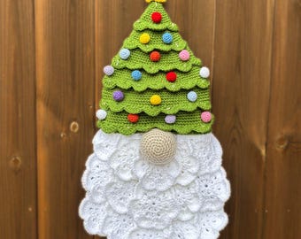 Op de afbeelding: Een handgemaakte gehaakte kerstkabouterdecoratie. De kabouter heeft een groene boomvormige hoed versierd met kleurrijke pompons en een gele ster. De baard is wit en de neus is een beige bol. De kabouter wordt getoond tegen een houten achtergrond.
