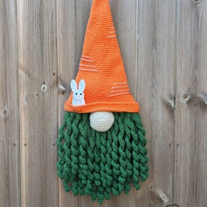 May include: A handmade crochet gnome with an orange carrot-shaped hat, white bunny applique, green beard, and a beige nose. The gnome is a decorative item, likely for Easter or spring.