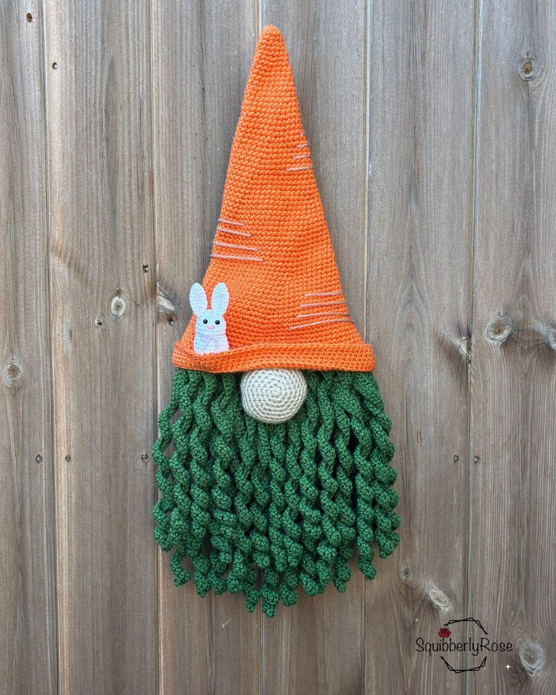 May include: A handmade crochet gnome with an orange carrot-shaped hat, white bunny applique, green beard, and a beige nose. The gnome is a decorative item, likely for Easter or spring.
