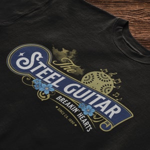 A balck t-shirt on a dark wooden background. The t-shirt has a vintage design (like 1800's soap advertisement) that says "Steel Guitar, breakin' hearts since 1890s"