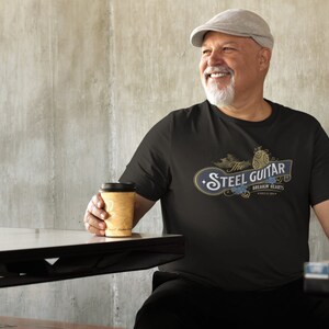 An older, heavy-set white male drinking coffee and wearing a black t-shirt. The t-shirt has a vintage design (like 1800's soap advertisement) that says "Steel Guitar, breakin' hearts since 1890s"