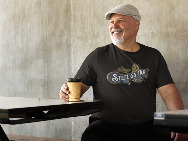 An older, heavy-set white male drinking coffee and wearing a black t-shirt. The t-shirt has a vintage design (like 1800's soap advertisement) that says "Steel Guitar, breakin' hearts since 1890s"