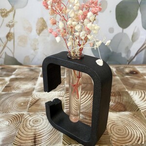 Peut inclure: Un vase décoratif noir en forme de U contenant un tube en verre transparent avec des fleurs séchées roses et blanches. Le vase est posé sur une surface en bois avec un motif circulaire. Les fleurs ont un aspect délicat et aérien.