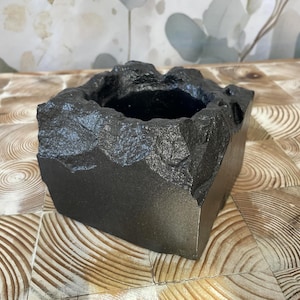 Peut inclure: Un pot carré noir avec un bord texturé en forme de roche. Le pot a un intérieur lisse et sombre et un extérieur uni et mat. Le design donne l'apparence d'un petit récipient en pierre, adapté à l'intérieur ou à l'extérieur.