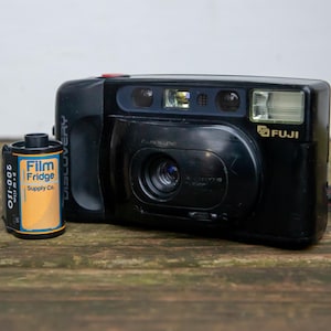 May include: Black Fuji Discovery camera with a roll of Film Fridge Supply Co. film. The camera has a red button and a built-in flash. The film canister is blue and orange.