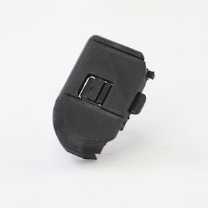 Pode incluir: Um objeto preto impresso em 3D, com formato retangular e um pequeno botão saliente. O objeto tem uma superfície texturizada e parece ser um componente de um dispositivo. O fundo é branco.