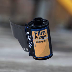 May include: A black and orange film roll with the text "Film Fridge Supply Co." and "200-ISO B+W film". The film roll is partially unrolled, revealing the film strip. The label has a blue border and a yellow background.