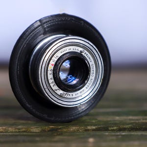 Op de afbeelding: Close-up van een vintage cameralens met een zwarte rubberen kap. De lens is zilverkleurig met zwarte markeringen en de tekst "MADE IN U.S.A.". De lens heeft een f/3.5 50 mm ARGUS COATED CINTAR markering.