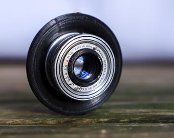 May include: Close-up of a vintage camera lens with a black rubber hood. The lens is silver with black markings and the text "MADE IN U.S.A." The lens has a f/3.5 50 mm ARGUS COATED CINTAR marking.