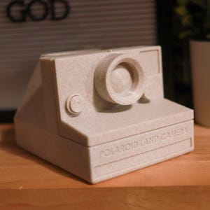 Könnte beinhalten: Eine weiße, aus Kunststoff gefertigte Nachbildung einer Polaroid Land Kamera. Die Kamera hat eine strukturierte Oberfläche und die Worte "POLAROID LAND CAMERA" sind auf der Vorderseite aufgedruckt.