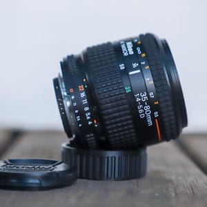 Op de afbeelding: Een zwarte Nikon AF Nikkor 35-80mm 1:4-5.6D cameralens met een rubberen scherpstelring met textuur, witte en oranje bedrukte tekst en een losse zwarte lensdop. De lens is voorzien van een metalen vatting en diverse diafragma-markeringen.