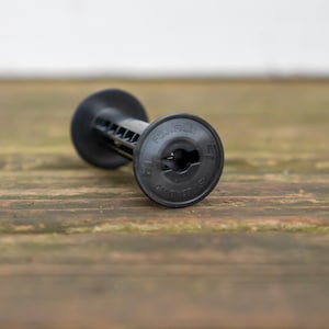 May include: A black Fujifilm film developing spool, lying on a wooden surface. The spool has a cylindrical shape with a central opening and the Fujifilm logo is visible. The spool is designed for 35mm film.