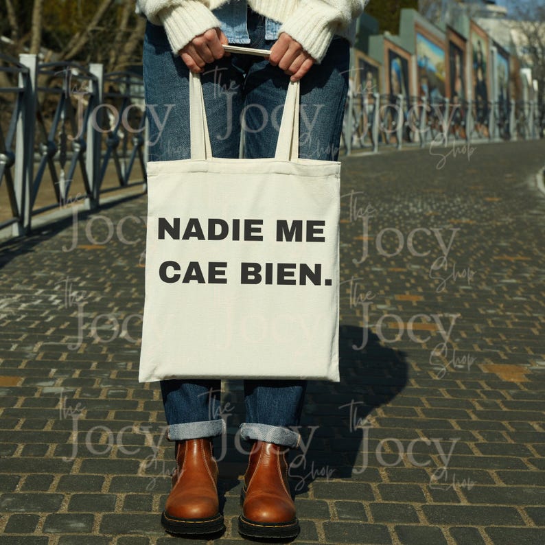 Puede incluir: Bolso tote de color crema con el texto en espa&ntilde;ol "NADIE ME CAE BIEN" en negro. El bolso lo sostiene una persona que lleva vaqueros azules y botas marrones. El fondo es un camino de ladrillos.