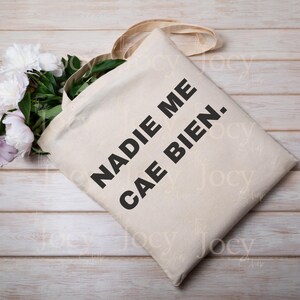 Puede incluir: Bolso tote beige con el texto "NADIE ME CAE BIEN." en letras negras en negrita. El bolso tiene un dise&ntilde;o sencillo con asas largas. Un ramo de peon&iacute;as blancas y rosas descansa junto al bolso sobre una superficie de madera clara.