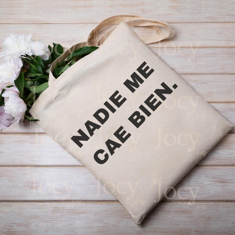 Puede incluir: Bolso tote beige con el texto "NADIE ME CAE BIEN." en letras negras en negrita. El bolso tiene un dise&ntilde;o sencillo con asas largas. Un ramo de peon&iacute;as blancas y rosas descansa junto al bolso sobre una superficie de madera clara.