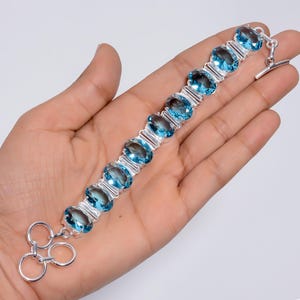 Puede incluir: Un brazalete de plata con piedras preciosas ovales de color azul claro. El brazalete se sostiene en una mano, mostrando las piedras pulidas y el cierre. Las piedras están engastadas en una banda de plata, creando un aspecto clásico y elegante.