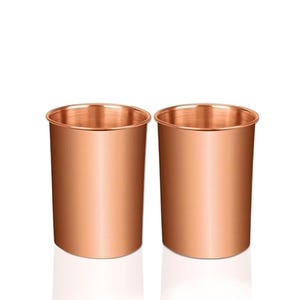 Könnte beinhalten: Zwei kupferfarbene Trinkgläser. Die Gläser sind zylindrisch mit einem leicht ausgestellten Rand. Das Kupfer hat einen warmen, metallischen Glanz. Die Gläser sind leer und stehen auf einer weißen Oberfläche. Die Gesamtästhetik ist schlicht und elegant.
