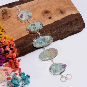 May include: A silver bracelet featuring five polished, oval-shaped stones in shades of mint green, teal, and deep magenta. The stones are set in smooth silver bezels and connected by small silver links, ending with a toggle clasp closure mechanism.