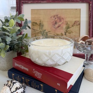 May include: A large, clear glass candle with a diamond pattern, filled with white wax. The candle sits atop two books with red and blue covers. A small potted plant and seashells are also in the image, along with a framed floral print.