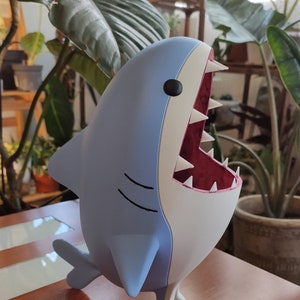 Peut inclure: Figurine de requin bleu clair et blanc avec une bouche ouverte révélant des dents blanches et un intérieur rouge. Le requin a des yeux noirs et des lignes courbes noires sur le côté. La figurine est sur une surface en bois.