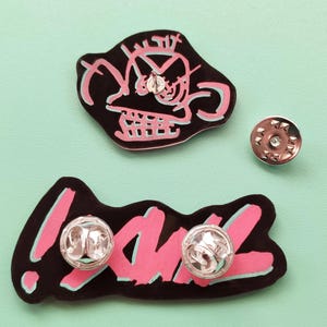 May include: Two enamel pins with black outlines and pink fill. One pin features a cartoon face with the text "!" and the other pin features the word "YAWL".